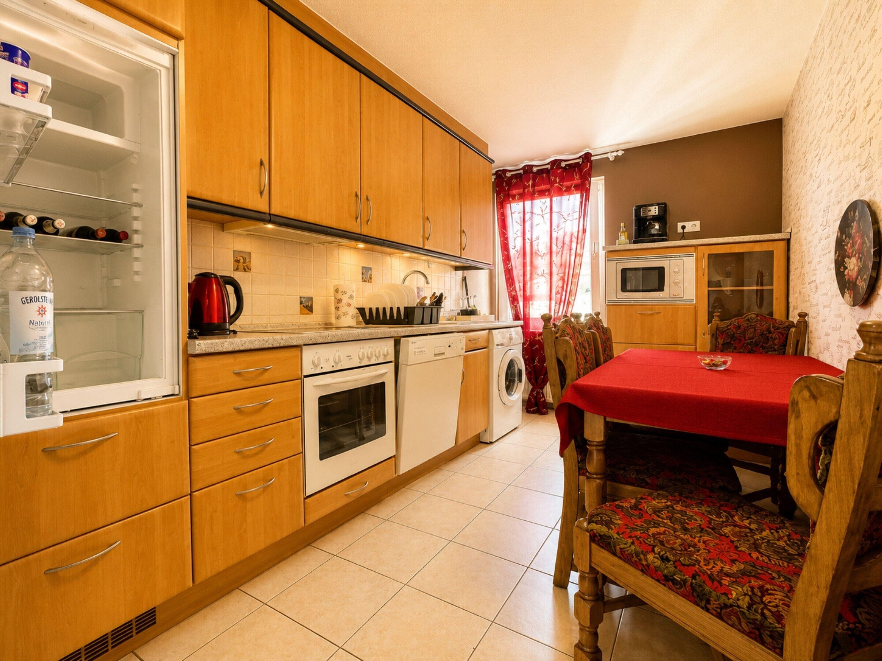 A compact kitchen with wooden cabinets, a built-in oven, a washing machine, and a dining area with a red tablecloth.