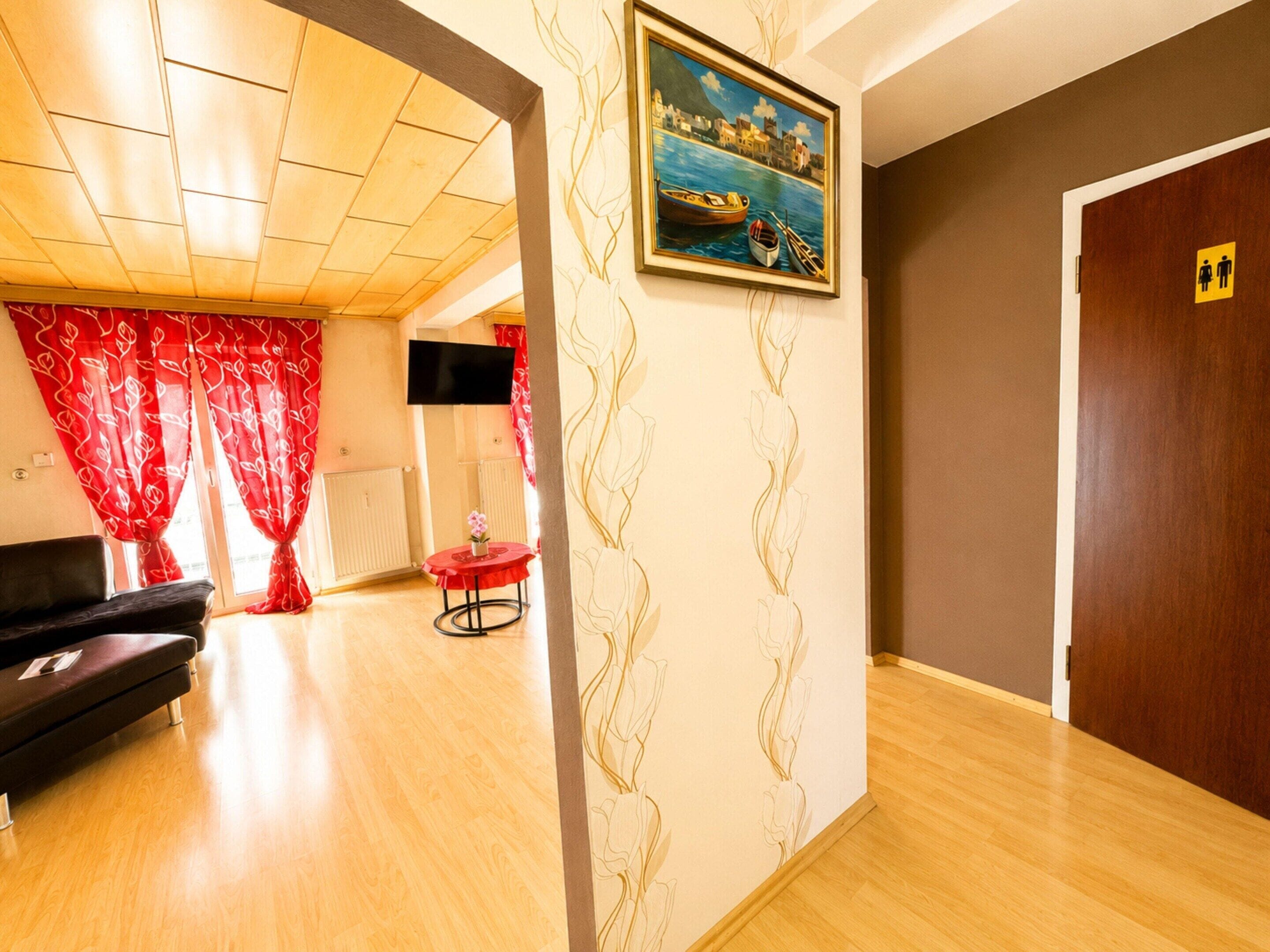 Ein Hotelzimmer mit Holzboden, einem schwarzen Sofa, einem roten Tisch mit einer Vase, einem Bild mit einer Küstenlandschaft und einer Tür mit der Kennzeichnung für Damen- und Herren-Toilette.