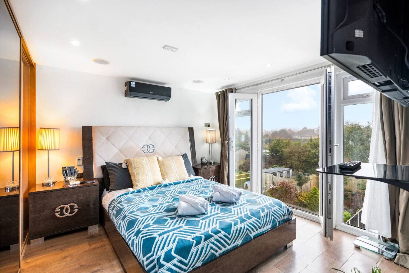 A modern bedroom with a large bed, a flat-screen TV, a window with a city view, and a built-in air conditioning unit.