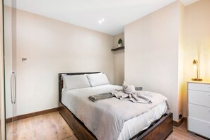 Ein Schlafzimmer mit Bett, Nachttisch, Kommode, einem Regal mit Pflanze und einer Wandleuchte.