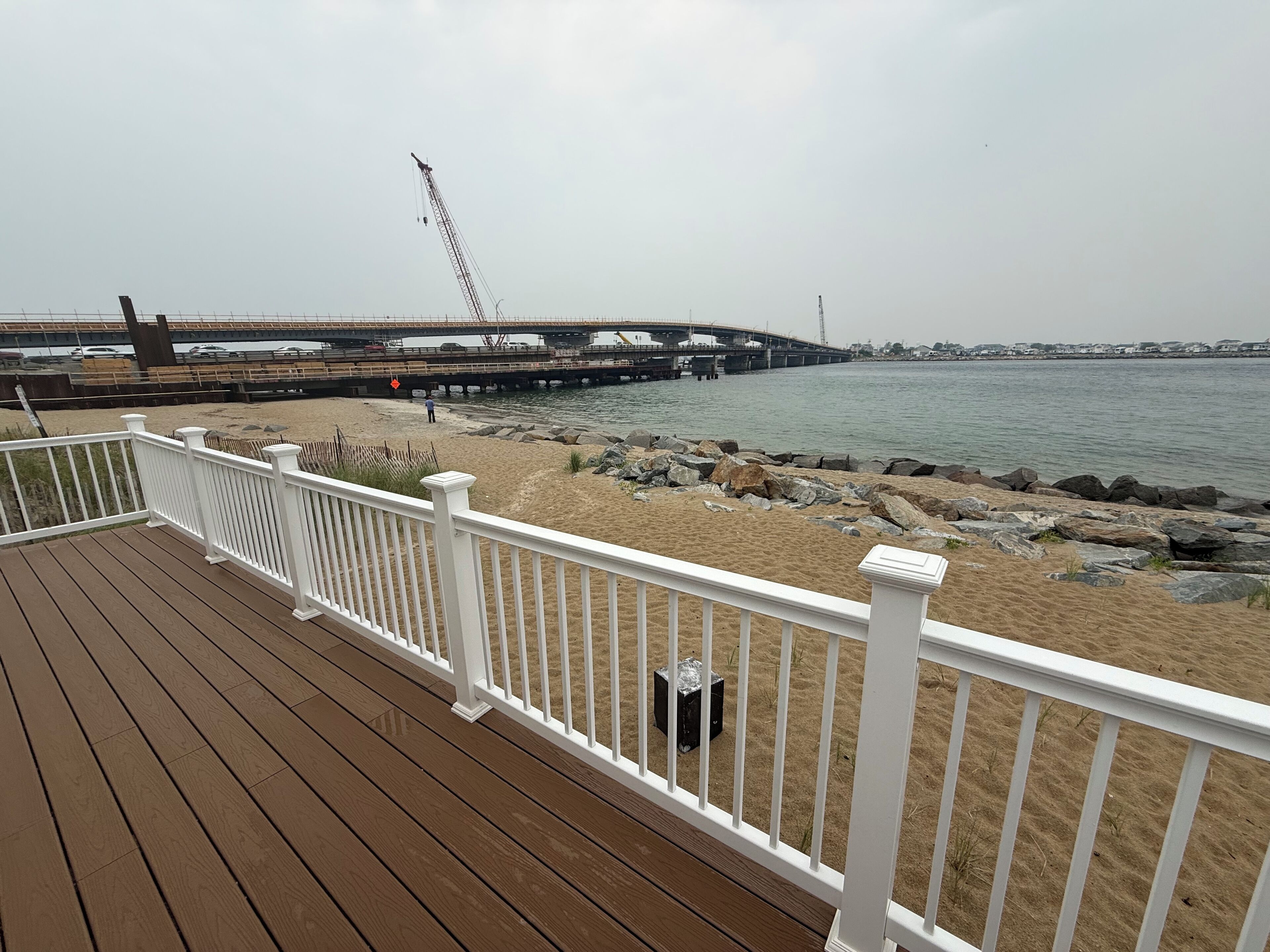 A wooden deck with white railings overlooking a beach with a pier and a bridge in the background.