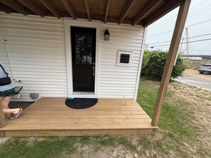 A wooden deck with a black door and a small window, surrounded by a grassy yard. - Driftwood House. Sleeps 4. Beach Access, Parking (Seabrook)