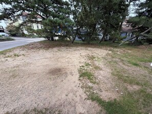A residential area with a gravel driveway, a lawn, and a large pine tree. - Driftwood House. Sleeps 4. Beach Access, Parking (Seabrook)