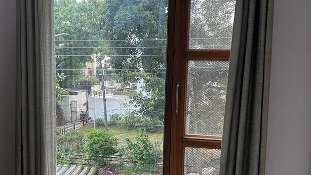A room with a window, a bed, and a view of a street with trees and parked vehicles. - Charming B&B Retreat in the Heart of Cha (Chandigarh)