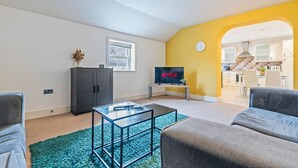 Uma sala de estar moderna com uma mesa de centro de vidro, sofás cinza, uma TV exibindo 'NETFLIX' e uma cozinha visível por meio de um arco.