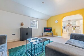 Ein modernes Wohnzimmer mit einem Glastisch, grauen Sofas, einem Fernseher, der 'NETFLIX' anzeigt, und einer durch einen Bogen sichtbaren Küche.