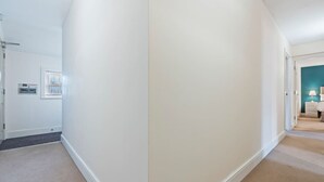 Um corredor com piso coberto por carpete, paredes brancas e uma porta que dá para um quarto com cama e mesa de cabeceira.