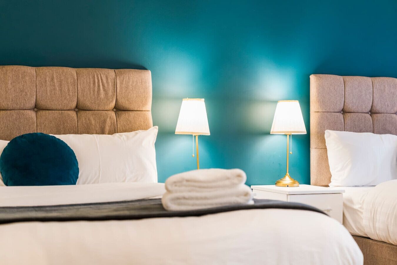 Et hotelværelse med to senge, hver med en polstret sengegavl, hvidt sengetøj og en teal-farvet væg.