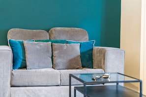 Ein graues Sofa mit türkisen und grauen Kissen vor einer türkisfarbenen Wand, davor ein schwarzer Tisch.