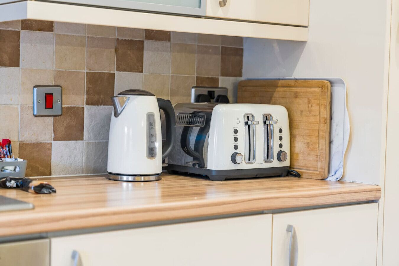 A kitchen counter with a kettle, toaster, and a cutting board.