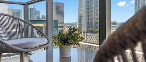 A balcony with a wicker chair, a small table with a plant, and a view of high-rise buildings.