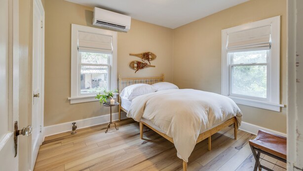 A bedroom with a bed, two windows, a bedside table with a plant, and an air conditioning unit on the wall.