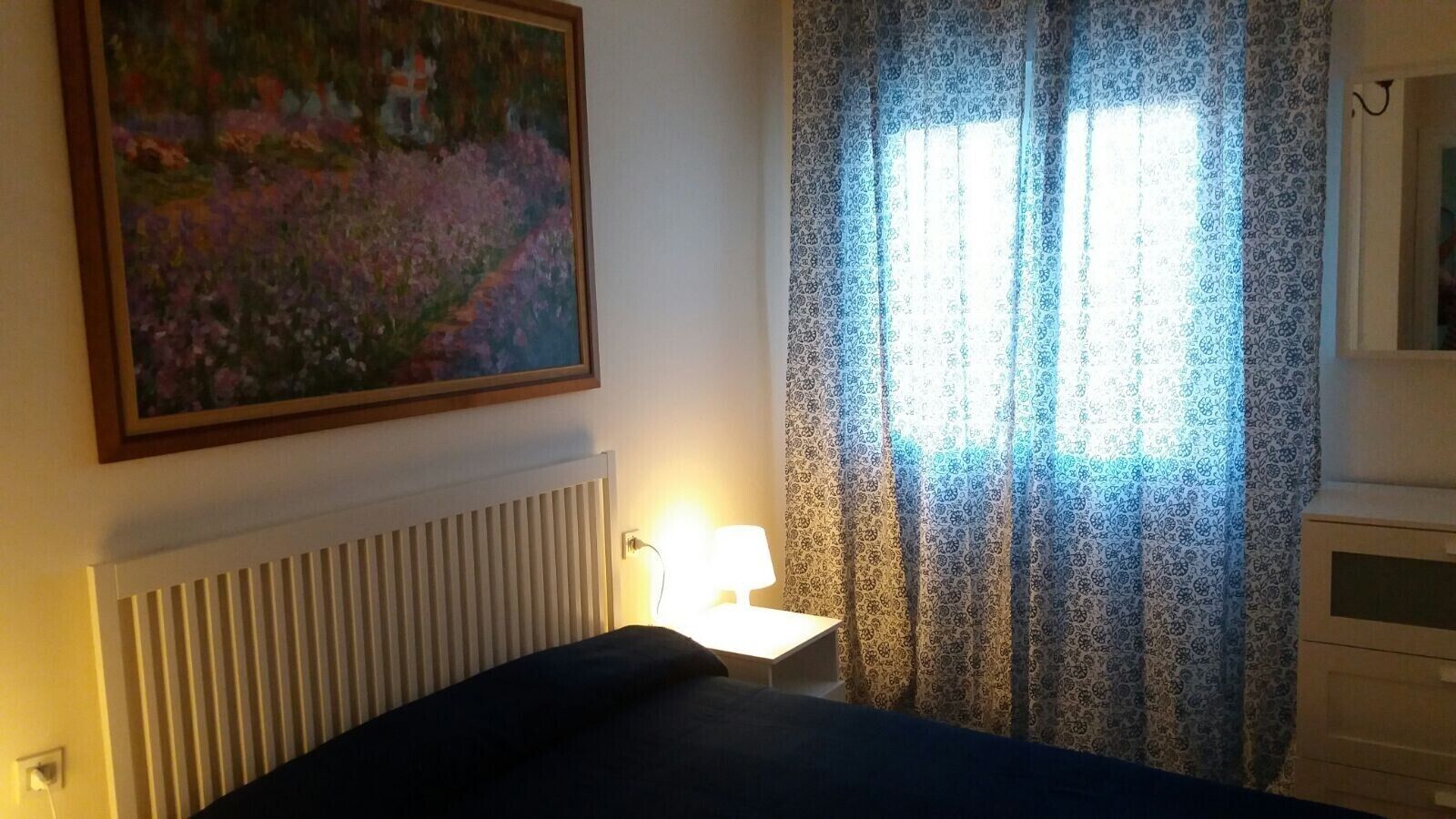 A bedroom with a bed, a bedside lamp, a radiator, a painting on the wall, and patterned curtains.