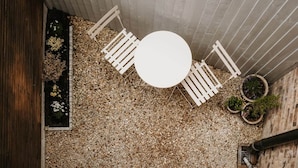 A top-down view of a patio area with a round table and white chairs, surrounded by gravel and potted plants.