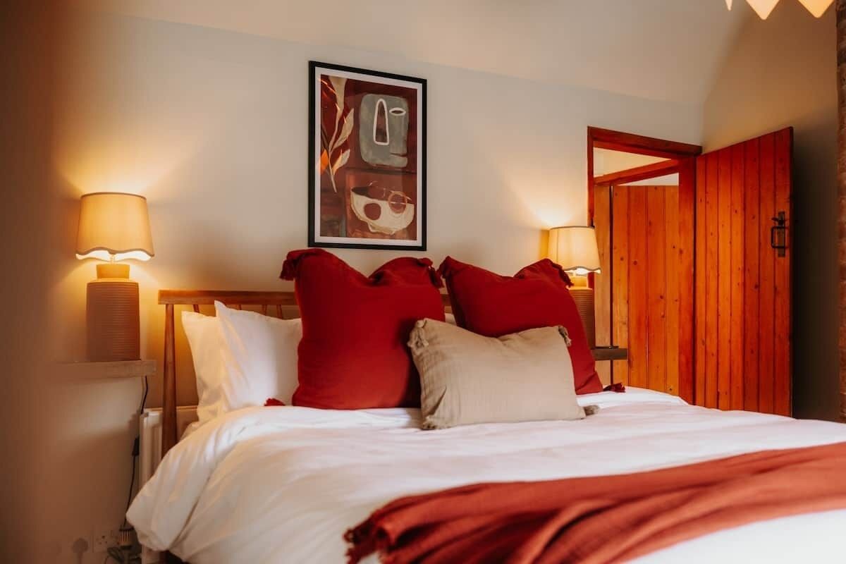 Ein Hotelzimmer mit Bett, roten und weißen Kissen, einem gerahmten Kunstwerk über dem Bett und einer Holztür.