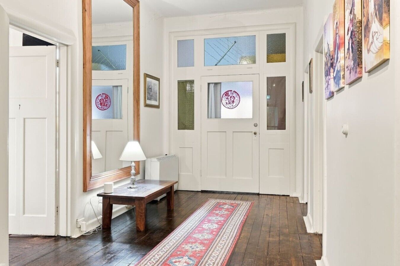 Ein Flur mit Holzboden, einer weißen Tür mit Fensterglas, einem Spiegel, einem Tisch mit Lampe und einem Teppich mit Muster.