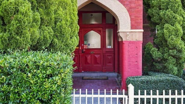 Un bâtiment en briques rouges, avec une porte rouge, entouré de haies bien entretenues et d’une clôture en bois peinte en blanc.