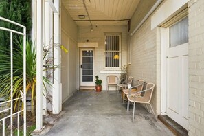 A narrow outdoor corridor with a concrete floor, a white door, and a window with bars. There are wicker chairs and a potted plant. - Edinburgh Family Room (Hobart)