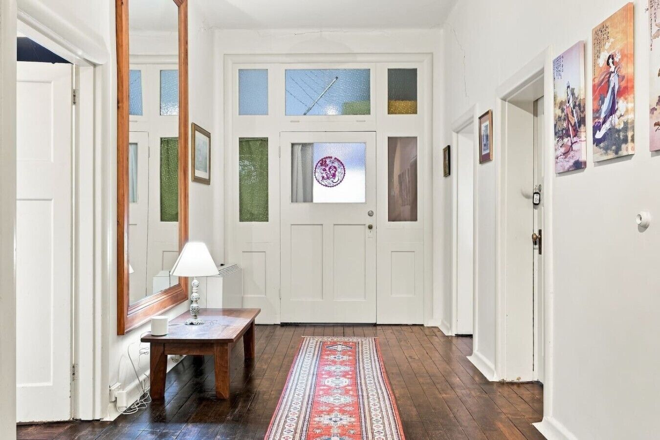 A hallway with a wooden floor, a white door with a frosted glass window, a small table with a lamp, and a patterned rug.