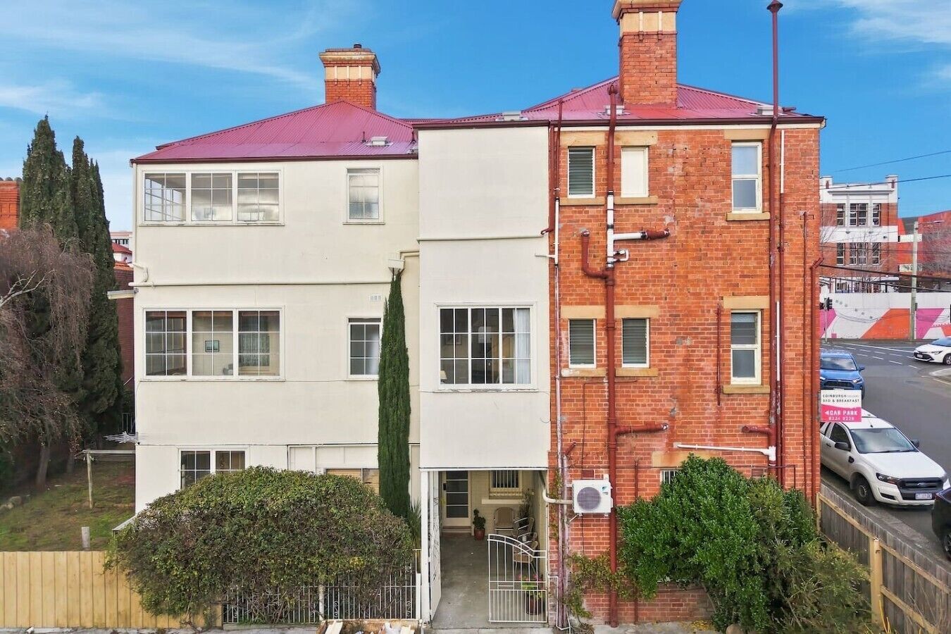A two-story building with a red roof, a white facade, and a brick section. There is a car parked in front, and a sign indicating a car park.