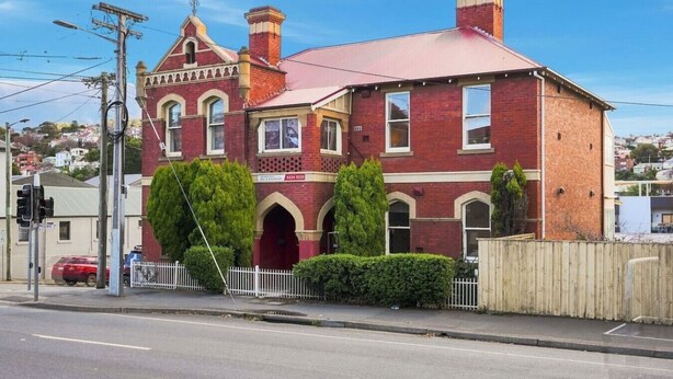 A two-story red brick building with a red roof, arched windows, and a white picket fence. - Edinburgh Queen & Single Rm + Ensuite (Hobart)