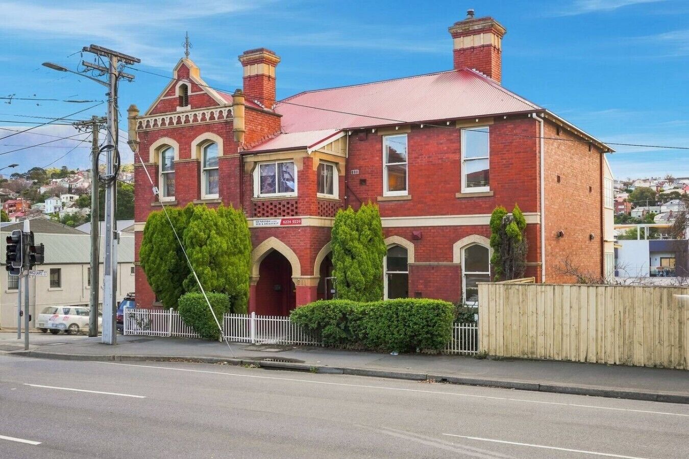 A red brick Victorian-style building with a prominent arched entrance, a fenced yard, and a clear sky.