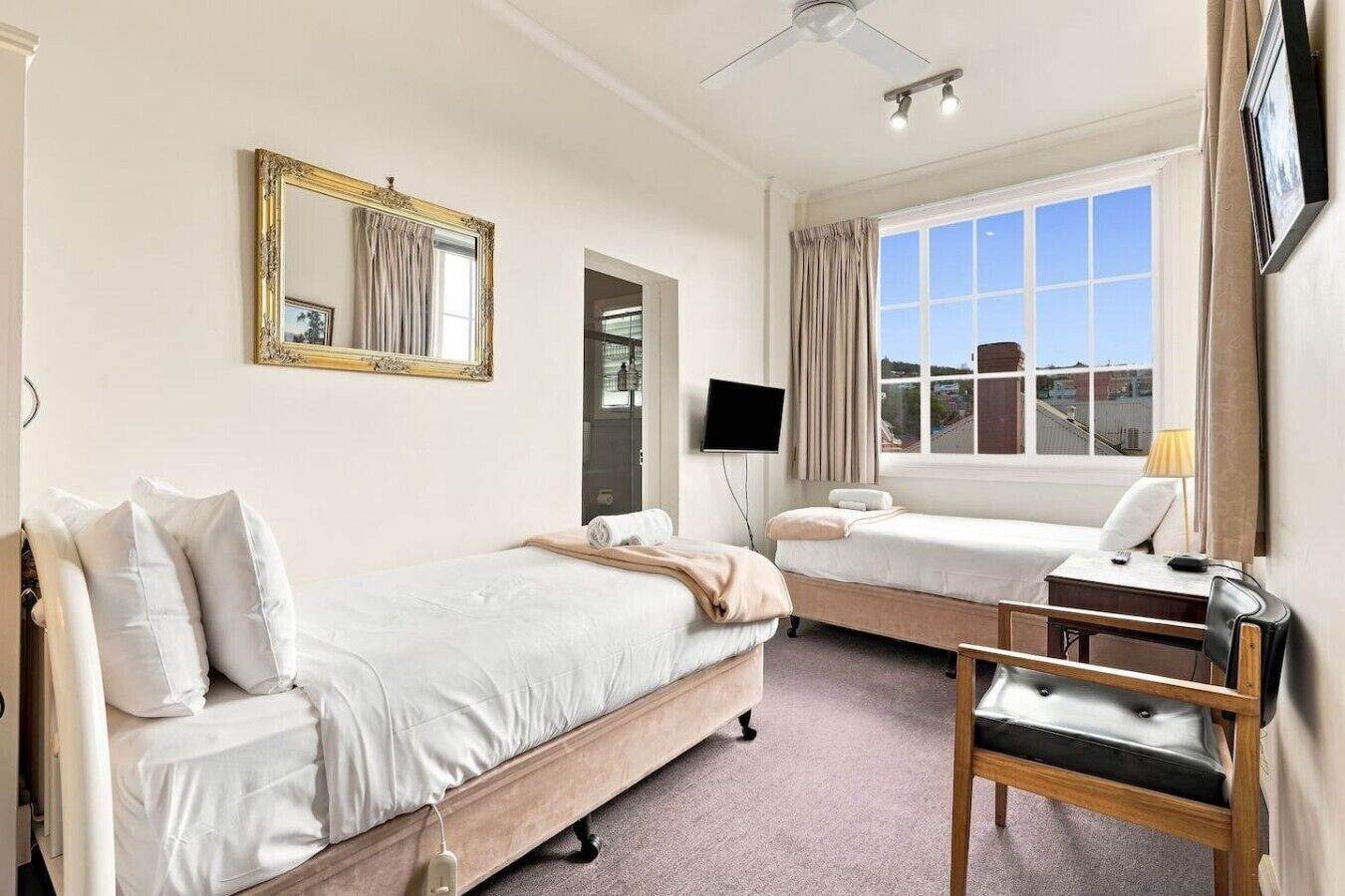 A hotel room with two beds, a desk, a chair, a television, a ceiling fan, and a large window with a view.