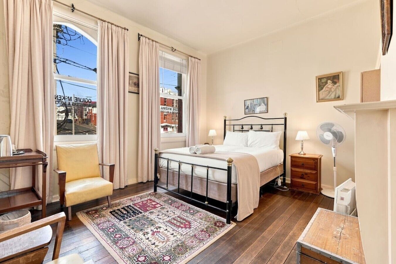 Un dormitorio con una cama, una silla, un escritorio, un ventilador de pie y una ventana con cortinas.