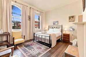 A bedroom with a bed, a chair, a desk, a standing fan, and a window with curtains. - Edinburgh Queen Room (Hobart)