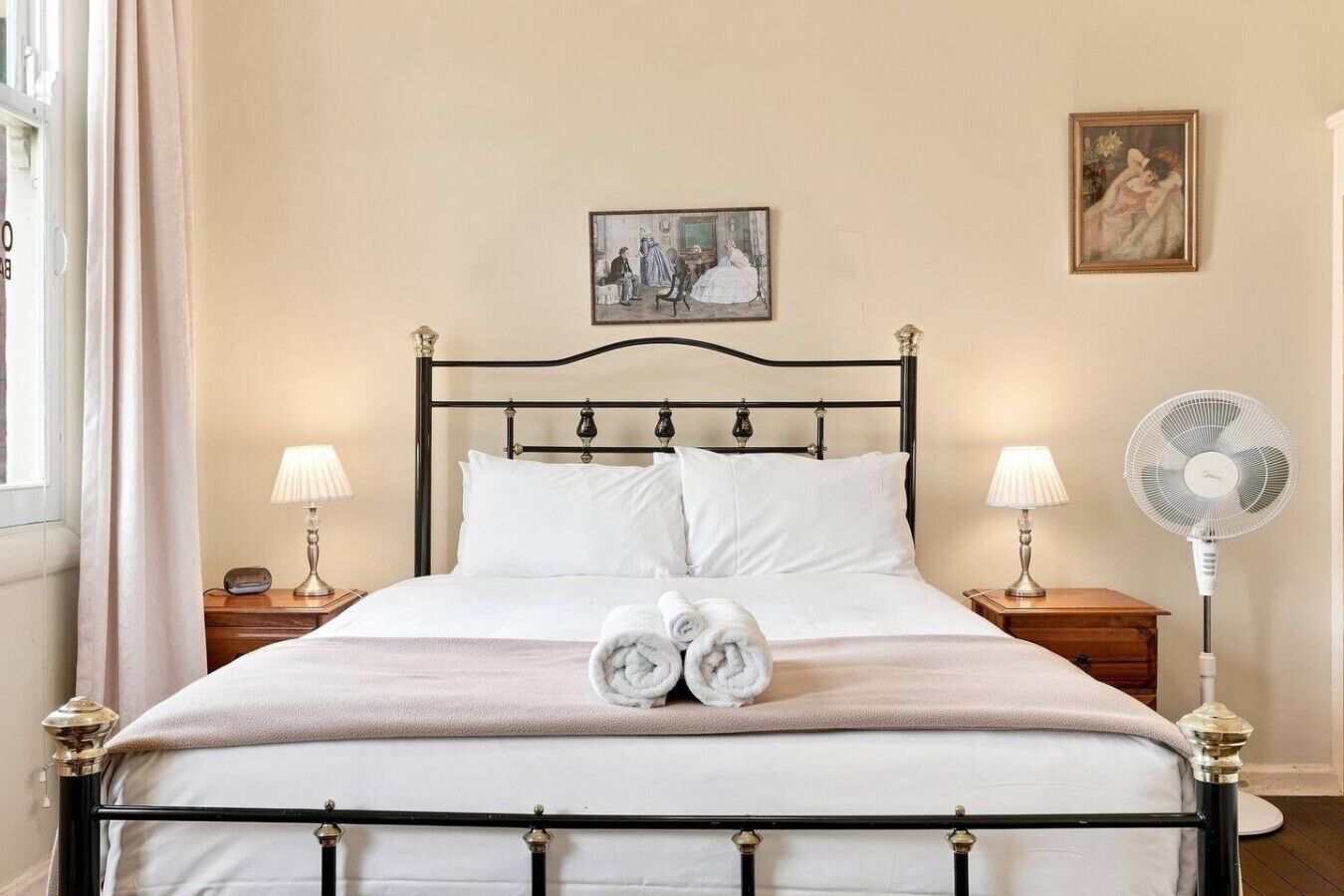 Un lit bien fait, avec du linge de lit blanc, deux lampes de chevet, un tableau accroché au mur et un ventilateur sur pied.