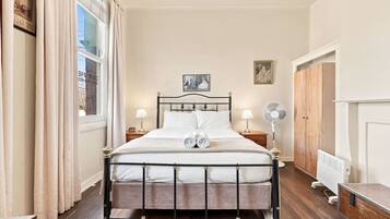 A neatly made bed with a headboard, two bedside lamps, a standing fan, and a radiator.