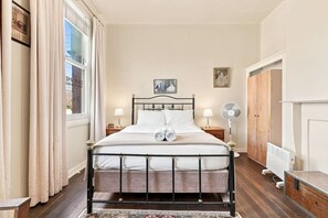 A neatly made bed with a headboard, two bedside lamps, a standing fan, and a radiator. - Edinburgh Queen Room (Hobart)
