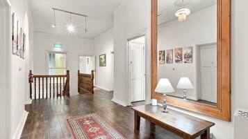 A hallway with a wooden floor, a red patterned rug, a bench with two lamps, and a large mirror on the wall.