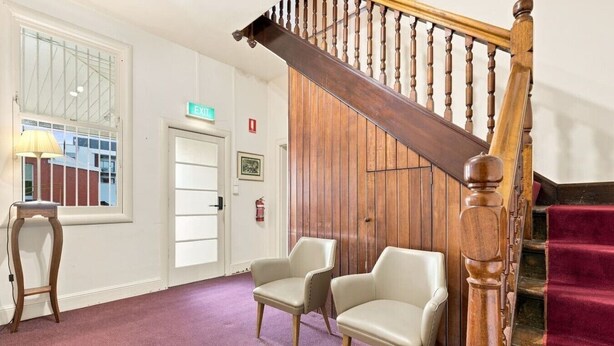 A hallway with a wooden staircase, two chairs, a lamp, and an exit sign. - Edinburgh Family + Ensuite | Hobart CBD (Hobart)
