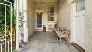 A narrow outdoor corridor with a concrete floor, a white door, and a window with bars. There are wicker chairs and a potted plant near the door. - Edinburgh Family + Ensuite | Hobart CBD (Hobart)