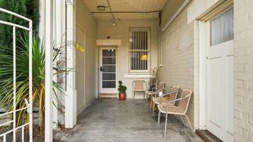 A narrow outdoor corridor with a concrete floor, a white door, and a window with bars. There are wicker chairs and a potted plant near the door.