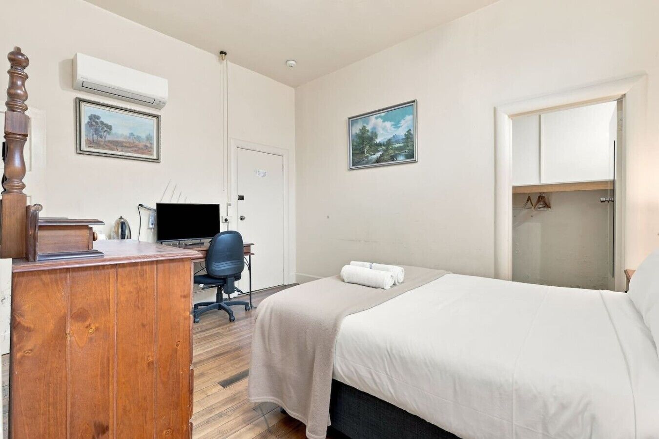 A hotel room with a wooden desk, a computer, a chair, two beds, a wardrobe, and two framed pictures on the wall.