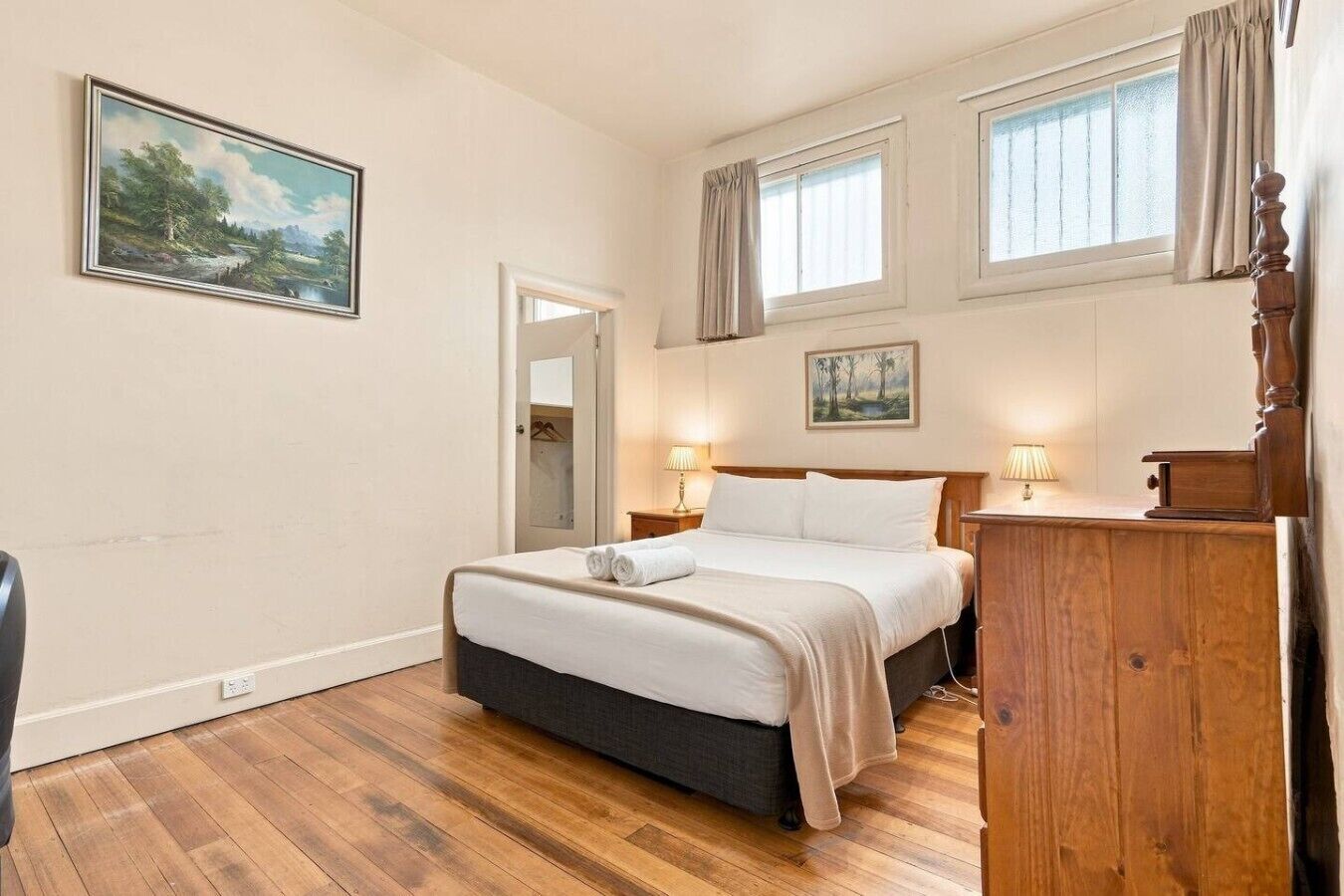 A bedroom with a bed, bedside lamps, a wooden dresser, a painting on the wall, and a window with curtains.