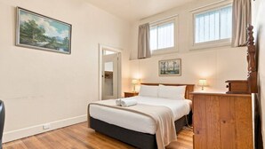 A bedroom with a bed, bedside lamps, a wooden dresser, a painting on the wall, and a window with curtains. - Edinburgh Family + Ensuite | Hobart CBD (Hobart)