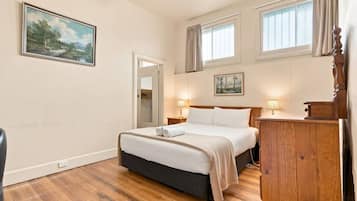 A bedroom with a bed, bedside lamps, a wooden dresser, a painting on the wall, and a window with curtains.