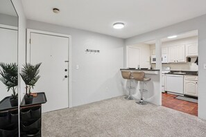 A modern kitchen with white cabinets, a microwave, and a coffee machine. There are two bar stools and a small table with a plant. - Cozy 1-BDR condo in Mansfield Close to commuter station to Boston & Providence (Mansfield)