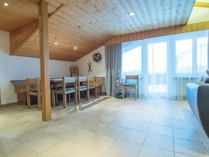 En rummelig stue med højt til loftet, træbjælker, et spiseområde med stole og bord, og et stort vindue med gardiner.
