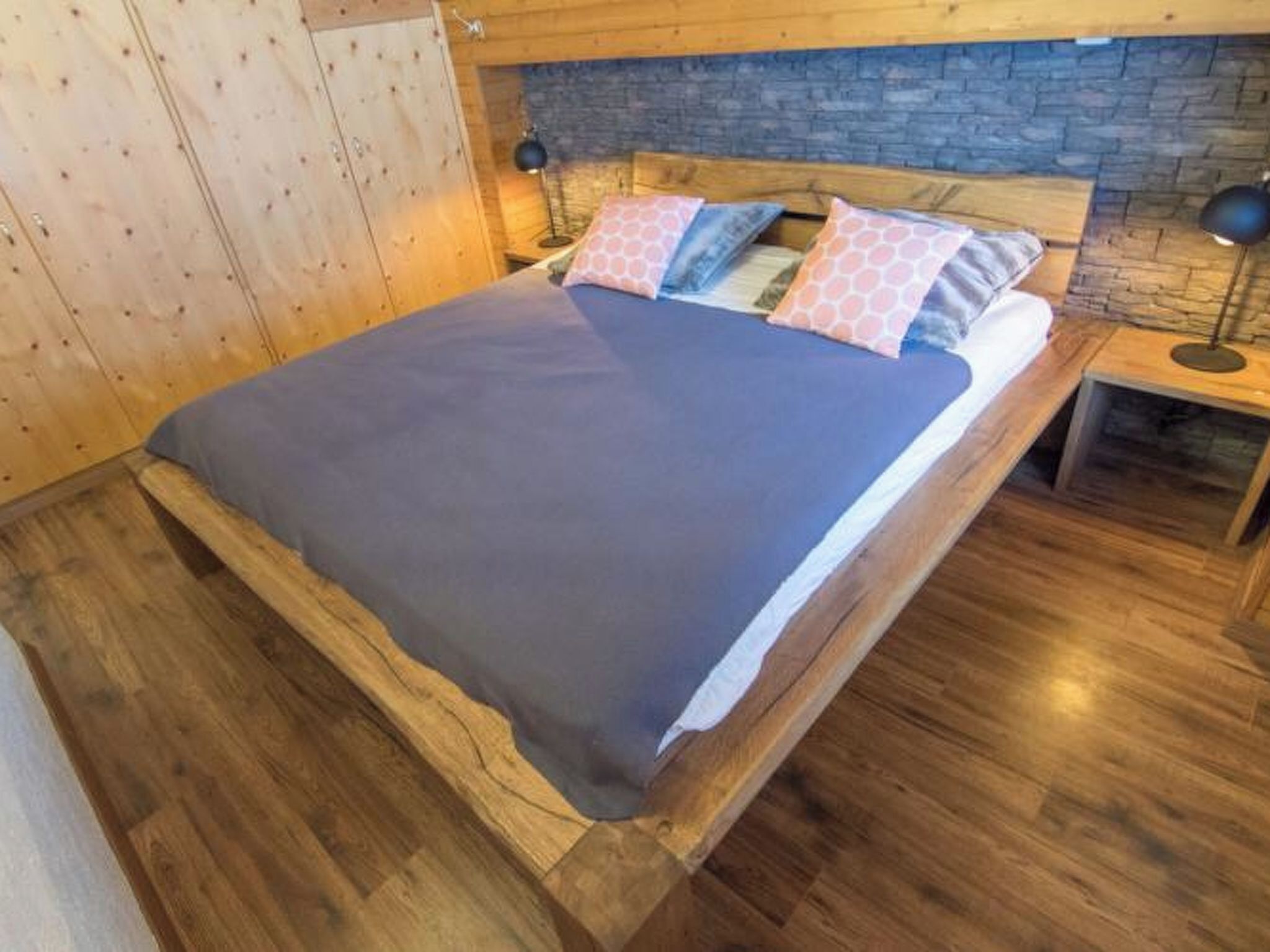 Un lit en bois, avec un mur en pierre en arrière-plan, une table de chevet et une lampe.