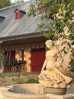 A stone statue of a woman seated on a pedestal, with a building featuring a red door and a cart in the background. - La Sellerie - Gîte de Charme à l'étage des écuries (Cour-Cheverny)