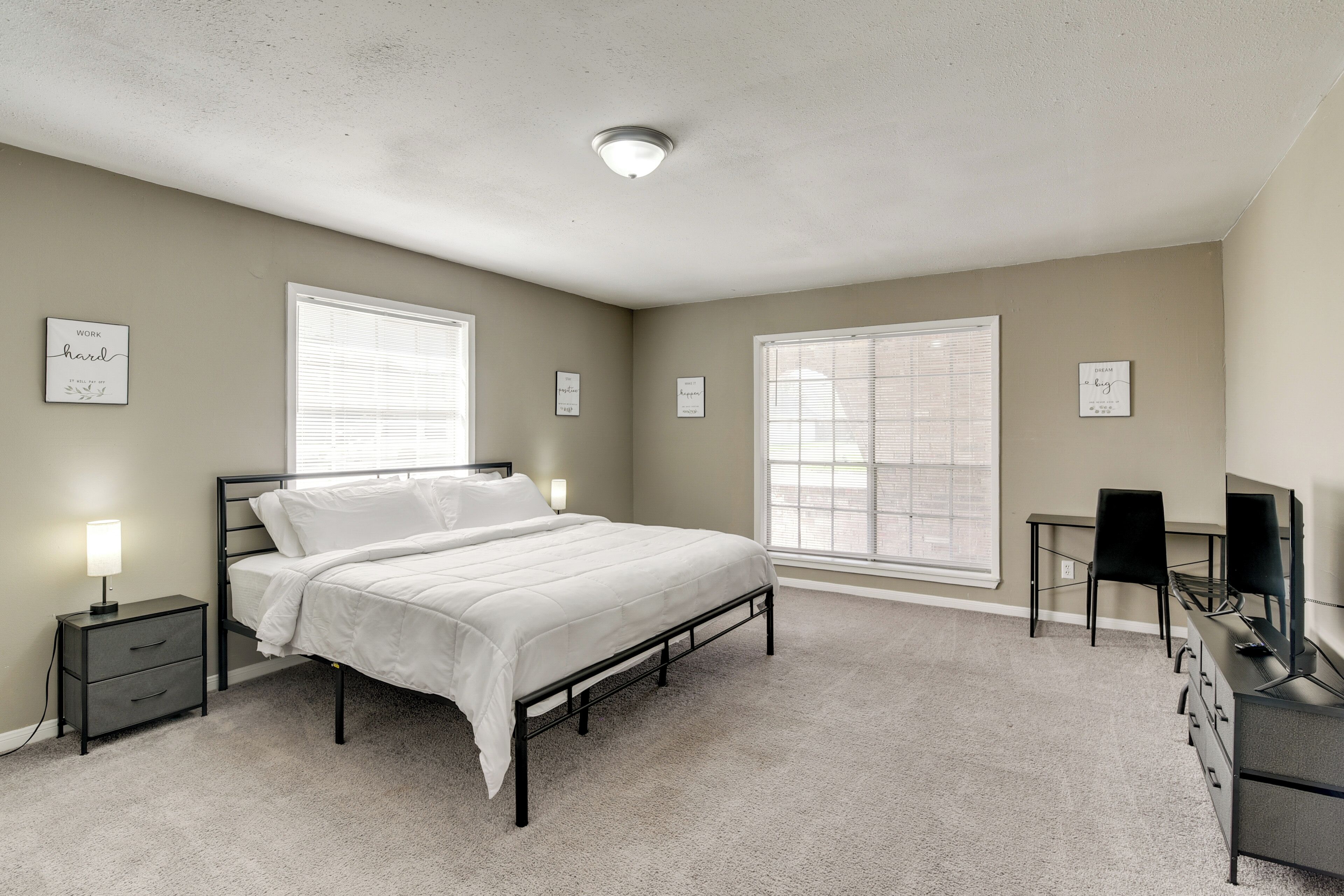 A bedroom with a bed, bedside tables, a desk with a chair, and large windows.