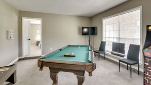 Una mesa de billar en una habitación con televisor, dos sillas y una ventana con persianas.