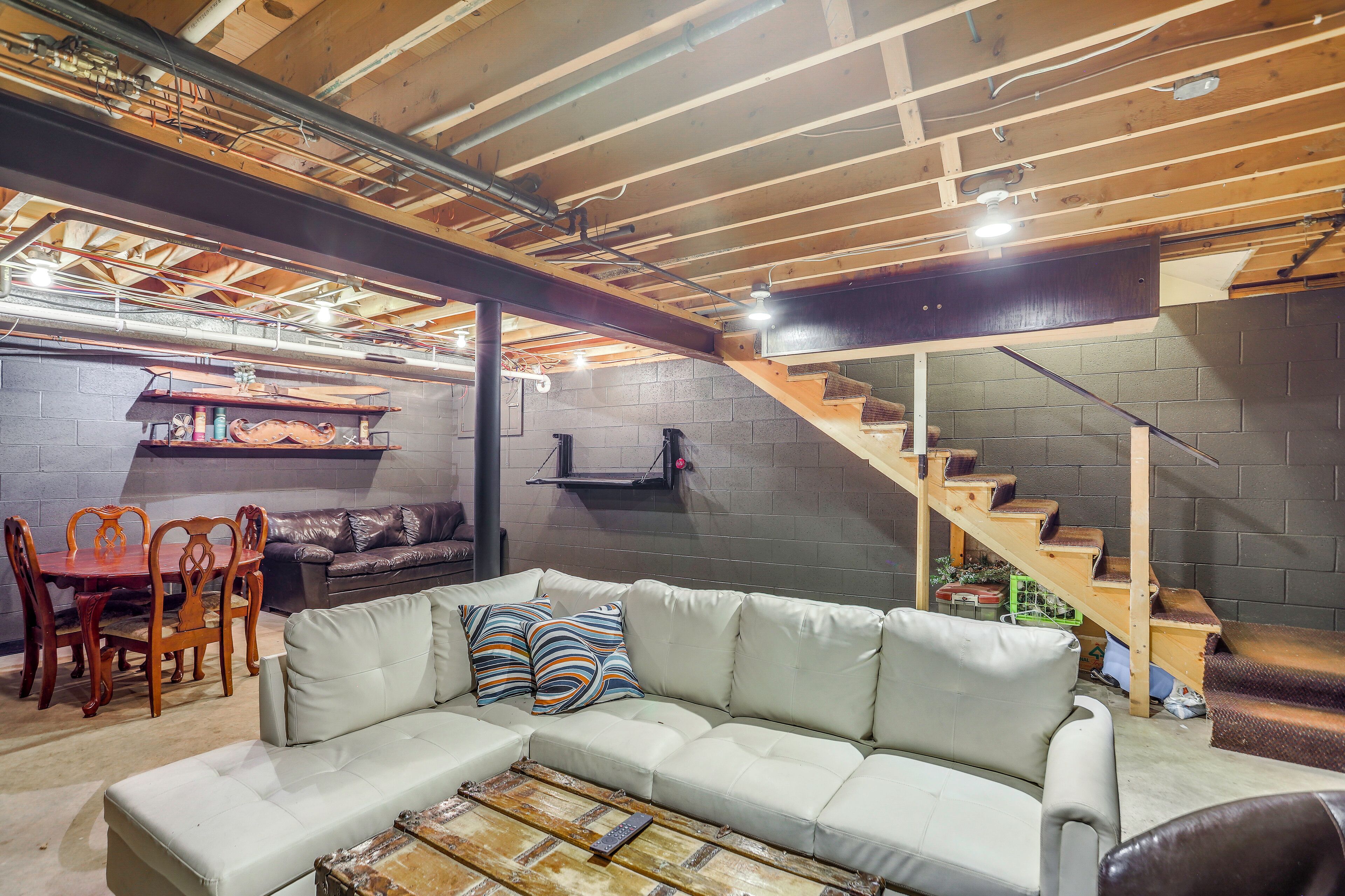 A cozy basement living area with a sectional sofa, a dining table with chairs, a staircase, and exposed wooden beams.