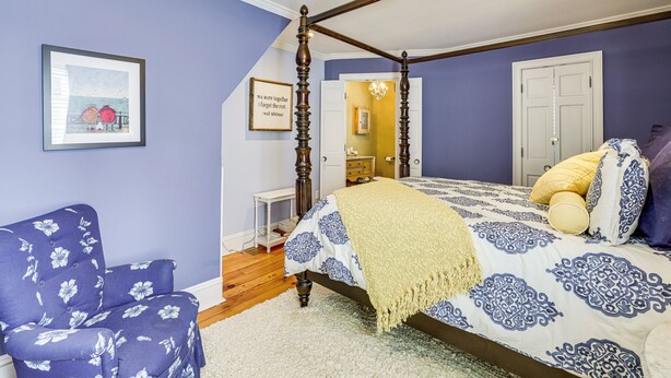 Un dormitorio con una cama con dosel, una silla, una cita enmarcada en la pared y una puerta que da a otra habitación.