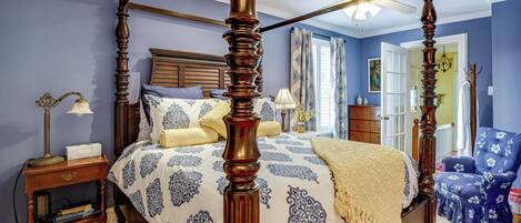 A four-poster bed with a canopy, a nightstand with a lamp, a chair with a floral pattern, and a wooden floor.
