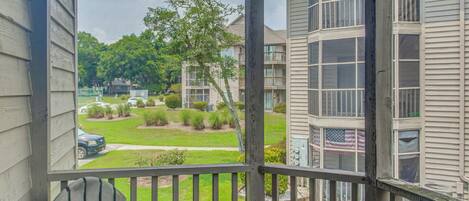A view from a balcony with a wooden railing, overlooking a residential area with houses and a lawn.